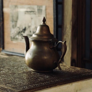 Könnte beinhalten: Eine antike Messingkanne mit einem verzierten Design steht auf einem weißen Tisch mit einer kunstvollen goldfarbenen Tischplatte. Der Tisch hat eine weiße, abgenutzte Oberfläche und gedrehte Beine.
