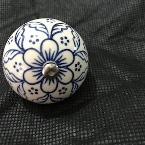 Könnte beinhalten: Ein weißer Keramik-Knopf mit einem blauen Blumenmuster. Der Knopf hat eine silberne Metallschraube in der Mitte.