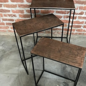 Könnte beinhalten: Drei Beistelltische mit rechteckigen, strukturierten, kupferfarbenen Oberflächen und schwarzen Metallrahmen. Die Tische sind unterschiedlich hoch, der höchste im Hintergrund. Die Tische stehen vor einer roten Backsteinmauer.