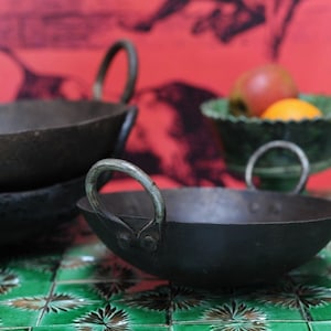 Könnte beinhalten: Drei schwarze Metallpfannen mit abgerundeten Griffen werden auf einer grün-weiß gefliesten Oberfläche präsentiert. Eine grüne Schüssel mit Obst befindet sich im Hintergrund vor einem roten Hintergrund. Die Pfannen haben ein rustikales, Vintage-Aussehen.