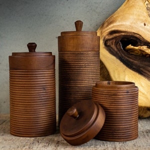 Könnte beinhalten: Vier Holzbehälter mit Deckeln in verschiedenen Größen. Die Behälter sind warmbraun und haben eine geriffelte Textur. Ein Deckel ist abgenommen und gibt den Blick ins Innere frei. Im Hintergrund befindet sich eine Holzplatte mit einzigartiger Maserung.