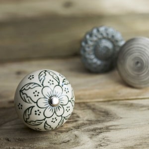 Könnte beinhalten: Drei Keramik-Knöpfe mit weißem Hintergrund und verschiedenen Designs. Ein Knopf hat ein Blumenmuster in Grün, ein anderer ein Wirbelmuster in Grau und der dritte ein Blumenmuster in Grau.