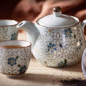 Könnte beinhalten: Ein Keramik-Teeservice mit einer Teekanne und zwei Tassen, alle mit einem Blumenmuster in Weiß, Blau und Braun verziert. Die Teekanne hat einen Deckel mit einem braunen Knauf und einem gebogenen Griff. Ein Tee-Ei aus Edelstahl ist ebenfalls vorhanden.