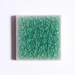 Könnte beinhalten: Eine quadratische Keramikfliese mit grüner Craquelé-Glasur. Die Fliese hat eine raue, strukturierte Oberfläche.