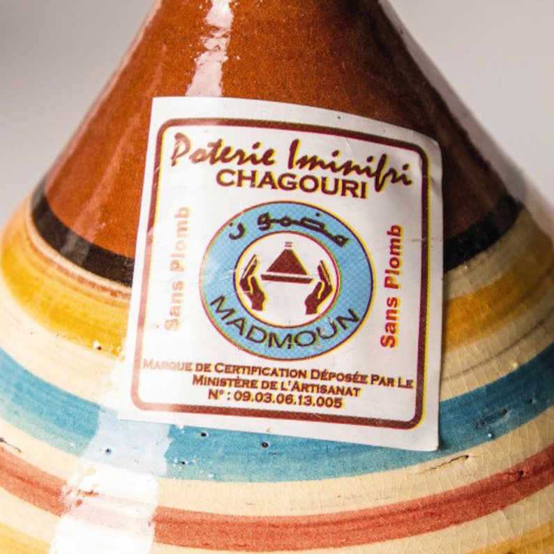 May include: A traditional Moroccan tagine, a conical clay pot with a colorful striped glaze. The tagine is labeled "Poterie Iminiri Chagouri Madmoun Sans Plomb" and has a certification mark from the Moroccan Ministry of Handicrafts.