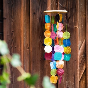 Könnte beinhalten: Ein farbenfrohes Windspiel mit einem hölzernen Oberteil und einer Reihe von hängenden, schillernden und durchscheinenden Formen in verschiedenen Farben. Die Formen sind meist rund oder quadratisch, und die Farben umfassen Blau, Rosa, Gelb und Orange. Der Hintergrund ist eine Holztür.