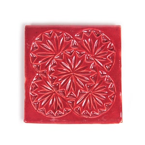 Könnte beinhalten: Eine rote Keramikfliese mit einem sich wiederholenden Muster aus vier geprägten, kreisförmigen Designs. Jedes Design zeigt ein Sternburst-Muster mit strahlenden Linien.