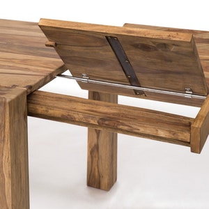 May include: A close-up of a wooden dining table with an extension leaf. The table is made of light brown wood and has a rectangular shape. The extension leaf is partially open, revealing the mechanism. The table legs are thick and square.