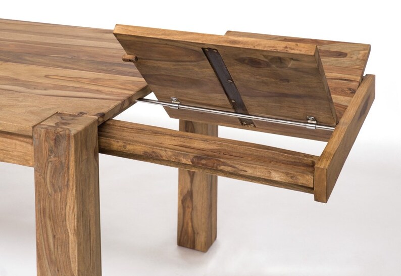 May include: A close-up of a wooden dining table with an extension leaf. The table is made of light brown wood and has a rectangular shape. The extension leaf is partially open, revealing the mechanism. The table legs are thick and square.