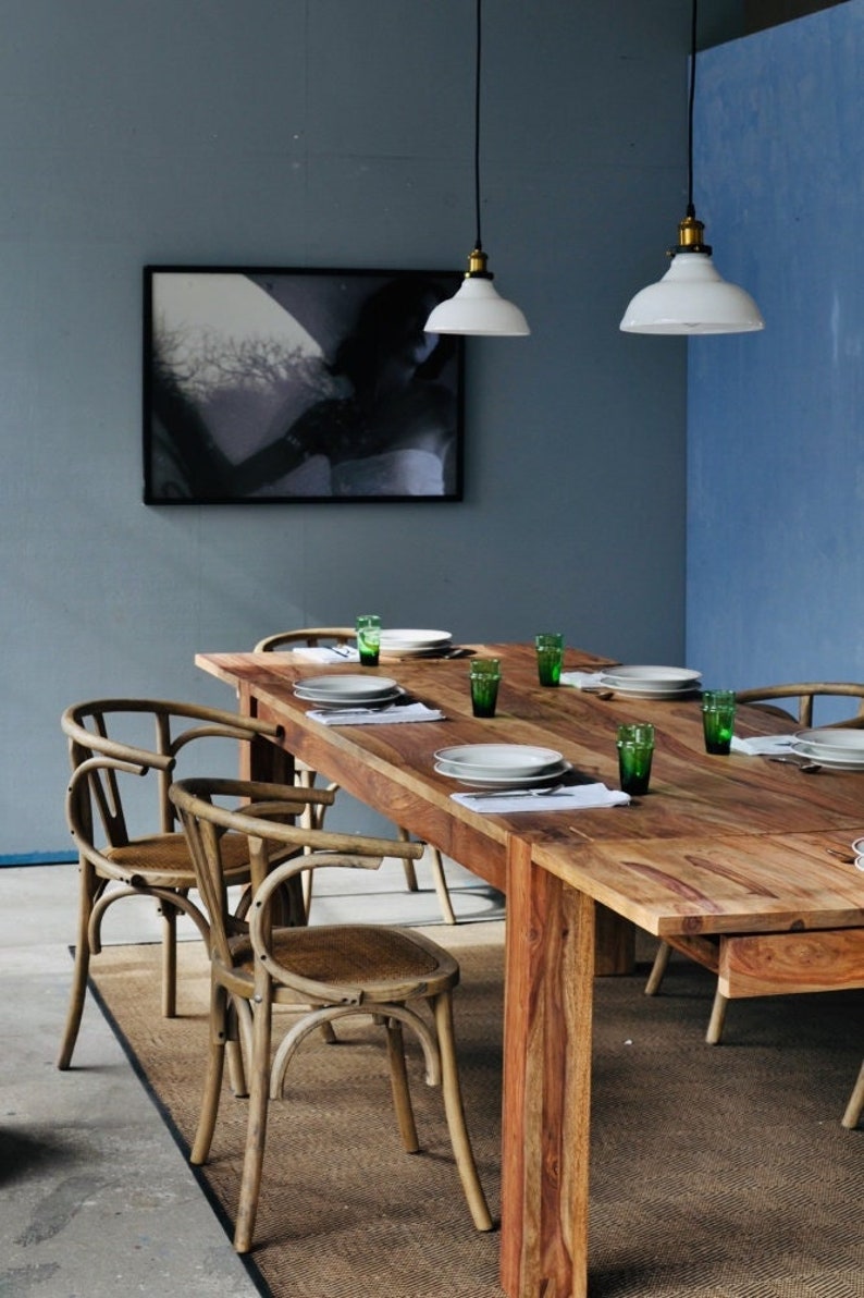 May include: A wooden dining table with six chairs, set with plates, napkins, and green glasses. Two white pendant lights hang above. A black and white framed artwork is on the wall.