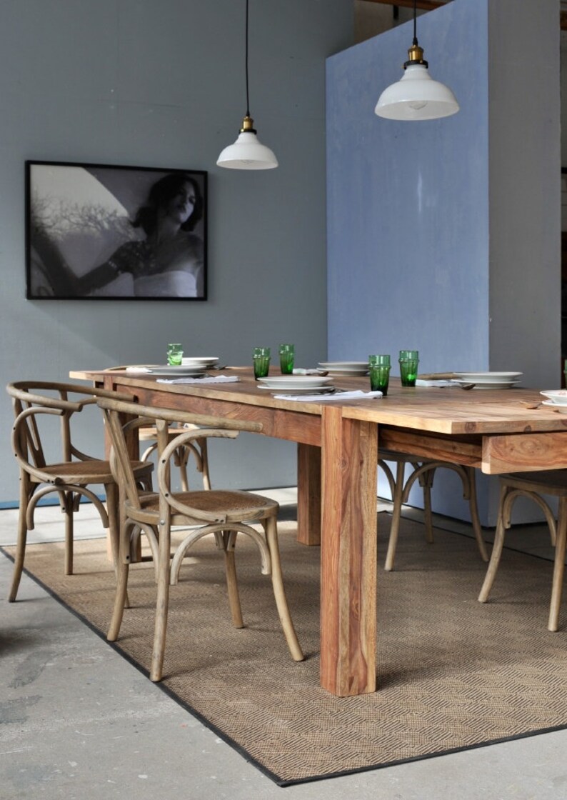 May include: A dining room scene featuring a large wooden table set with white plates and green glasses. Curved-back wooden chairs surround the table, and a framed black and white photograph hangs on the wall. Two pendant lights hang above the table.