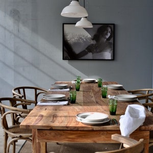 May include: A wooden dining table set for a meal, featuring plates, green glasses, and white napkins. The table is surrounded by wooden chairs. A black and white framed picture hangs on the wall, and two white pendant lights hang above the table.