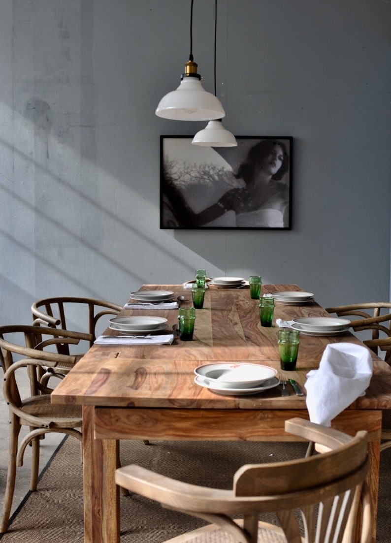 May include: A wooden dining table set for a meal, featuring plates, green glasses, and white napkins. The table is surrounded by wooden chairs. A black and white framed picture hangs on the wall, and two white pendant lights hang above the table.