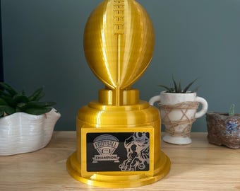 May include: A gold-colored football trophy with a detailed football shape on top. The trophy has a rectangular base with the words "FANTASY FOOTBALL CHAMPION" and a graphic of a football player. A decorative item for sports enthusiasts.