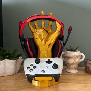 May include: A gold 3D printed hand holding a pair of red and black gaming headphones and a white Xbox controller. The hand is on a gold base.
