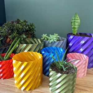 May include: A collection of 9 colorful, spiral-shaped plant pots in various colors, including red, yellow, blue, green, purple, and pink. The pots are made of a textured plastic material and are arranged on a wooden surface.