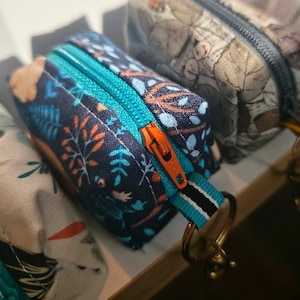 May include: Close-up of a small, rectangular zippered pouch with a teal zipper and orange pull. The pouch is navy blue with a botanical pattern in orange, teal, and white. Other pouches are visible in the background.