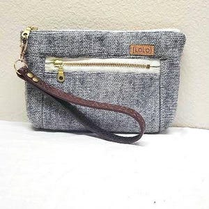May include: A gray and white fabric clutch with a brown leather strap and a zipper closure. The clutch has a small brown leather tag with the word "LOLO" on it.