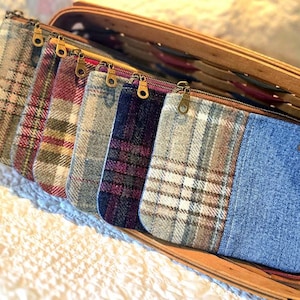 May include: A collection of rectangular zippered pouches arranged in a wooden basket. Each pouch features a unique combination of plaid wool fabric and blue denim, secured with a brass-toned metal zipper pull and a small rectangular leather tag.