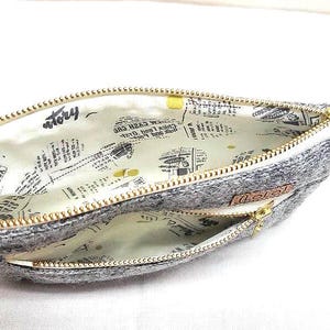May include: A gray zippered pouch with a double zipper closure. The pouch is lined with a white fabric with a newspaper print pattern. The text "story" is visible on the lining.