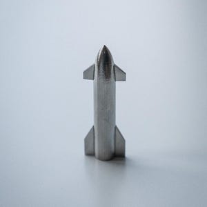 Pode incluir: Modelo de foguete prateado com um nariz pontudo e estruturas em forma de barbatana. O foguete tem um corpo cilíndrico e está sobre um fundo cinza claro. O modelo é uma representação em miniatura de uma nave espacial.