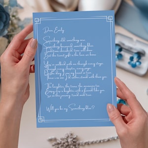 May include: A light blue card with white script, held by hands. The card has a decorative border and the text "Dear Emily" followed by a poem. The card is part of a wedding theme, with blue flowers and jewelry in the background.