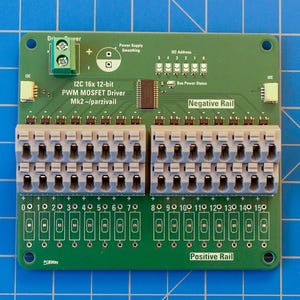 Może przedstawiać: Zielona płytka drukowana z oznaczeniem I2C 16x 12-bit PWM MOSFET Driver Mk2 ~/parzivail. Posiada zielony zacisk śrubowy, dwa białe złącza, centralny układ scalony oraz dwa rzędy szarych bloków zaciskowych z szesnastoma wyjściami.