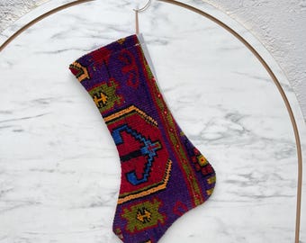 May include: A holiday stocking crafted from a vintage-style woven rug material featuring a vibrant geometric pattern in shades of deep purple, bright red, blue, green, and yellow. The stocking has a traditional boot shape with a hanging loop.