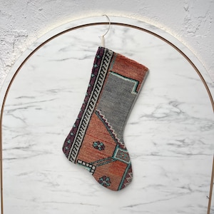 May include: A Christmas stocking crafted from vintage rug material featuring a geometric pattern in shades of orange, grey, and deep purple. The stocking has a textured, woven appearance with intricate border details and small floral motifs.
