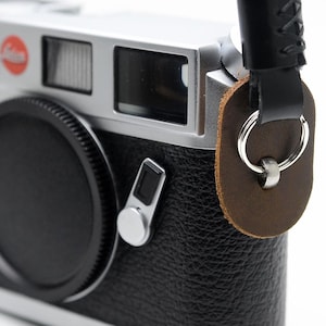 May include: A black and silver camera with a black lens cap and a black leather strap. The camera has a red dot and the word "Leica" printed on the top. The strap is attached to the camera with a brown leather piece and a silver ring.