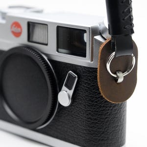 May include: A classic black and silver rangefinder camera with a black leather strap. The camera has a black lens cap and a red and white logo. The strap is attached with brown leather and silver rings.