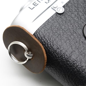 Może przedstawiać: Srebrno-czarny aparat Leica M6 z brązowym skórzanym mocowaniem paska. Aparat ma czarną osłonę obiektywu i czerwone logo Leica. Tekst "LEICA M6" jest widoczny na górze aparatu.