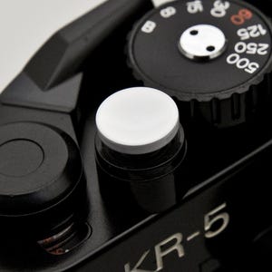 May include: Close-up of a black camera with a white button and a dial with numbers. The dial has numbers in white and red. The camera body is black and has the text "KR-5" in white.