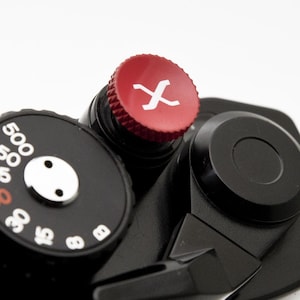 Pode incluir: Close-up de um corpo de câmera preto com um botão vermelho marcado com um "X" branco. Um mostrador preto com números brancos e um ponto branco também é visível. A câmera está em um fundo branco.