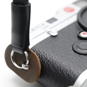 May include: A black and silver Leica camera with a black lens cap and a black leather strap. The camera has a textured black body and a silver base. The strap is attached to the camera with a brown leather piece and a metal ring.
