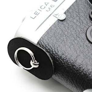 May include: A close-up shot of a Leica M6 camera, featuring a silver body and black leather grip. The camera has a red Leica logo and a black lens cap. The strap attachment includes a silver ring.