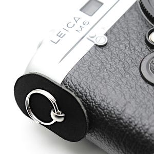 May include: Close-up of a silver and black Leica M6 camera. The camera has a black leather grip, a silver body, and a red Leica logo. The lens cap is black, and the camera strap attachment has a silver ring.