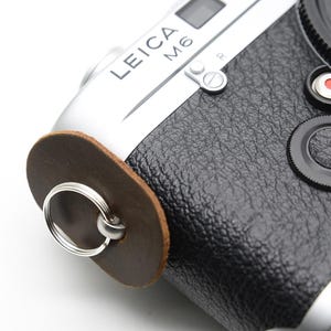 Może przedstawiać: Srebrno-czarny aparat Leica M6 z brązowym skórzanym mocowaniem paska. Aparat ma czarną osłonę obiektywu i czerwone logo Leica. Tekst "LEICA M6" jest widoczny na górze aparatu.