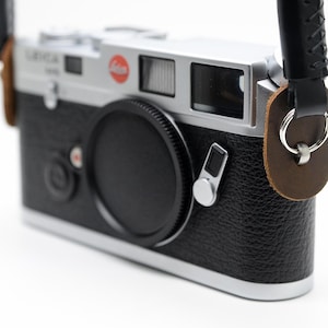 May include: A black and silver camera with a black lens cap and a black leather strap. The camera has a red circular logo and a silver top. The strap is attached with brown leather and silver rings.
