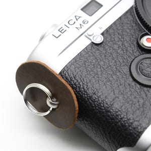 May include: A silver and black Leica M6 camera with a brown leather strap attachment. The camera has a black leather grip, a lens cap, and a red Leica logo. The text "LEICA M6" is visible on the top.
