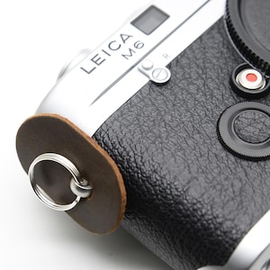 May include: A silver and black Leica M6 camera with a brown leather strap attachment. The camera features a black leather grip, a lens cap, and a red Leica logo. The strap attachment has a silver ring.