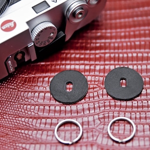 Może przedstawiać: Zbliżenie srebrnego aparatu fotograficznego z czarnymi skórzanymi akcesoriami i srebrnymi brelokami do kluczy na czerwonej, teksturowanej powierzchni. Aparat ma srebrną tarczę z numerami. Skórzane akcesoria są okrągłe z prostokątnym otworem pośrodku.