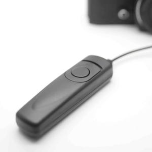 May include: A black remote shutter release with a single button, connected to a camera by a black cord.