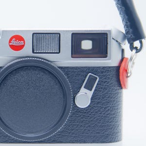 May include: A silver and black Leica M6 camera with a black lens cap and a black leather strap. The camera has a red Leica logo and red accents on the strap attachments. The camera is a classic film camera.