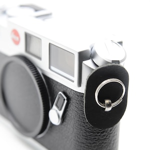Może przedstawiać: Srebrno-czarny aparat dalmierzowy z czarnym skórzanym etui i osłoną obiektywu. Aparat posiada logo z czerwoną kropką i srebrną metalową górną płytę. Aparat jest ustawiony pod kątem w prawo.