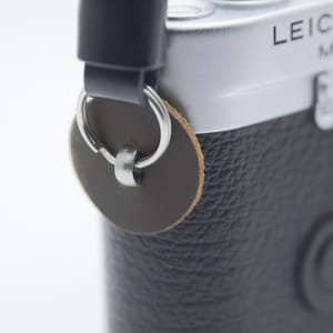 Può includere: Fotocamera Leica M6 argentata con impugnatura e copriobiettivo in pelle nera. Una tracolla nera è attaccata con un connettore in pelle marrone e anello argentato. Il logo Leica è visibile.