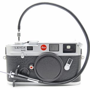 May include: A silver and black Leica M6 camera with a black lens cap and a black cable attached. The Leica logo is visible in red. The camera is on a white background.