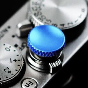 Könnte beinhalten: Nahaufnahme einer silbernen Kamera mit einem blauen Knopf. Der Knopf hat einen strukturierten Rand und ist von einem silbernen Ring umgeben.