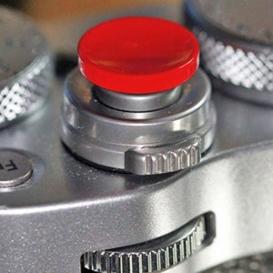 Op de afbeelding: Een close-up van een zilveren camera met een rode knop. De knop is gemarkeerd met "F" en is omgeven door een zilveren ring.