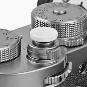 Könnte beinhalten: Nahaufnahme einer silbernen Kamera mit einem weißen Knopf und mehreren Drehknöpfen. Die Drehknöpfe sind mit Zahlen und Symbolen beschriftet, darunter "0", "1", "2", "3", "+", "-", "ISO", "250", "500", "1000", "2000", "4000", "8000", "125", "60", "30", "15", "8", "4", "2", "1" und "T".